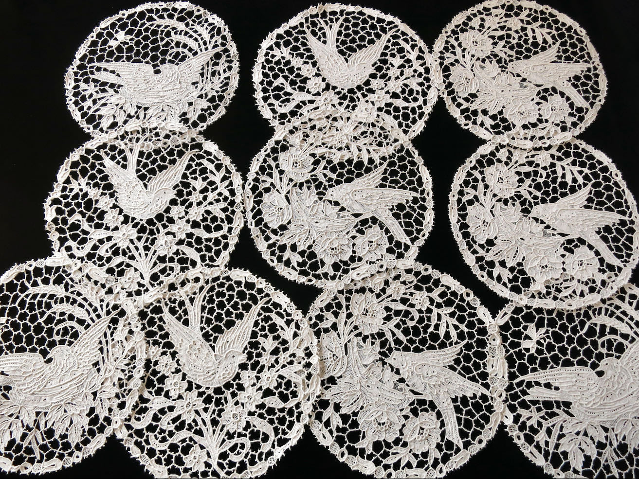 Collection of ten Italian handmade needle lace cocktail rounds, with bird scenes, laid out on a neutral background.