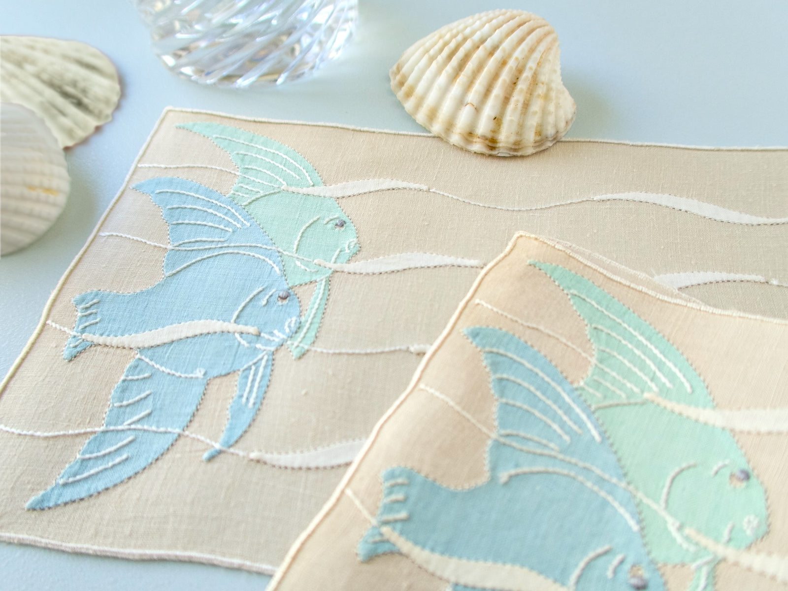  Close view of blue angelfish appliqué and embroidery on vintage Madeira linen cocktail napkin, undulating wave detail.​​​​​​​​​​​​​​​​