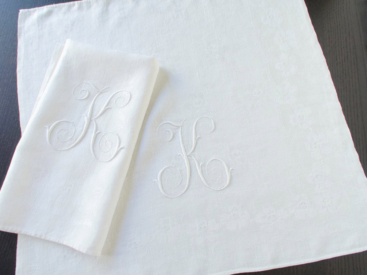 Folded and unfolded white damask linen napkins showing their large embroidered monograms.