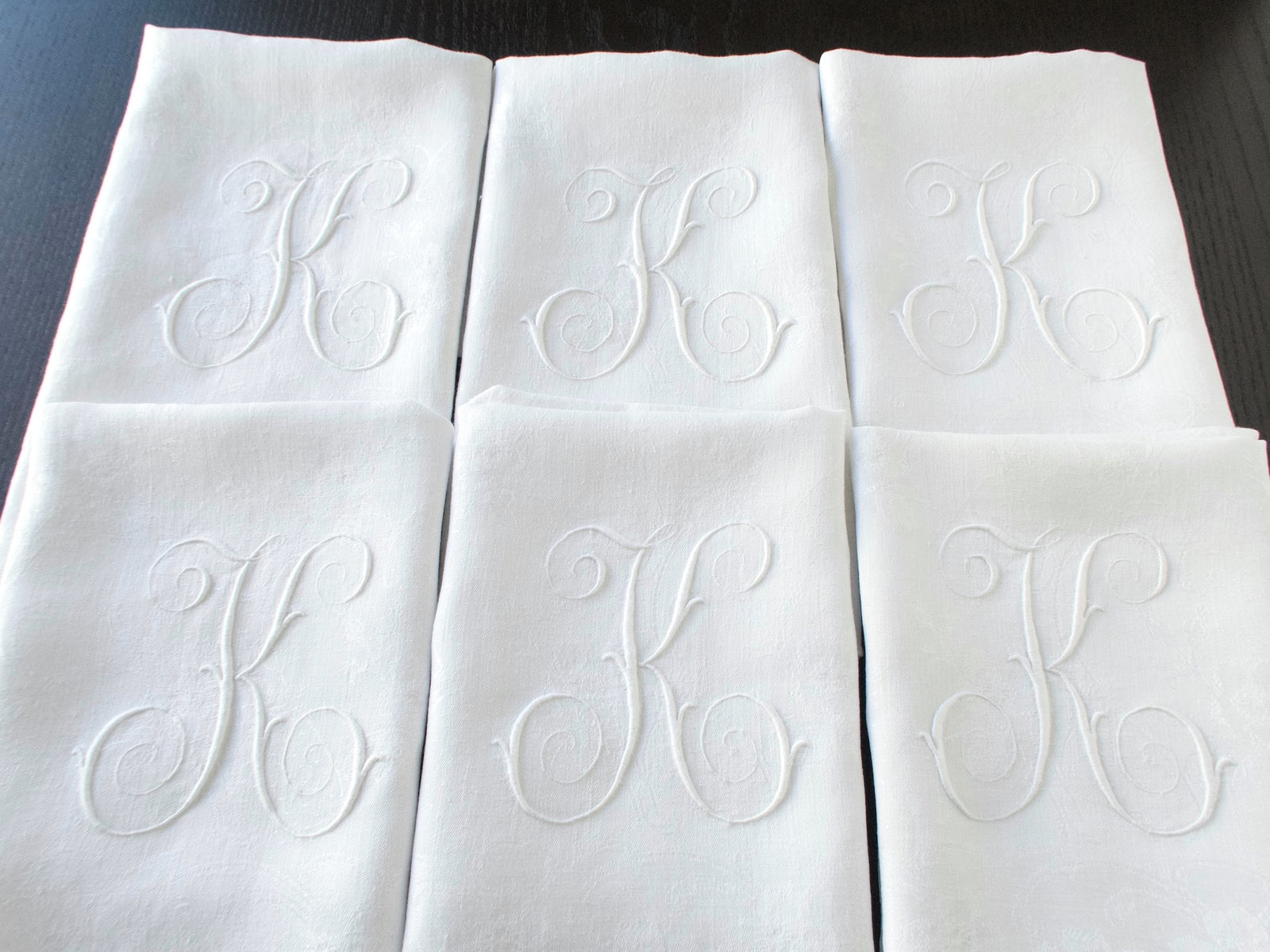 Six white damask linen napkins arranged to show their matching embroidered monograms.