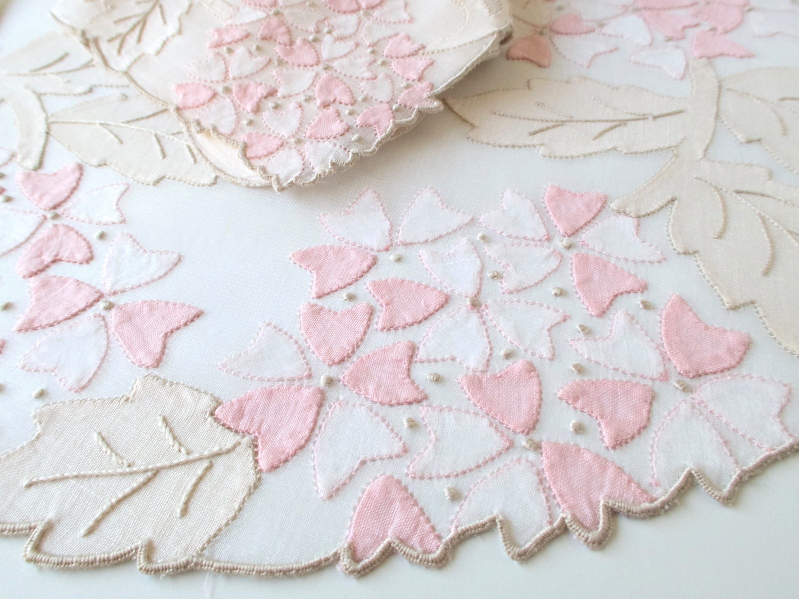 Detail of vintage Madeira organdy placemat with pale pink viburnum appliqué and hand embroidered flowers and ecru leaves on sheer ground, scalloped edge, white background.