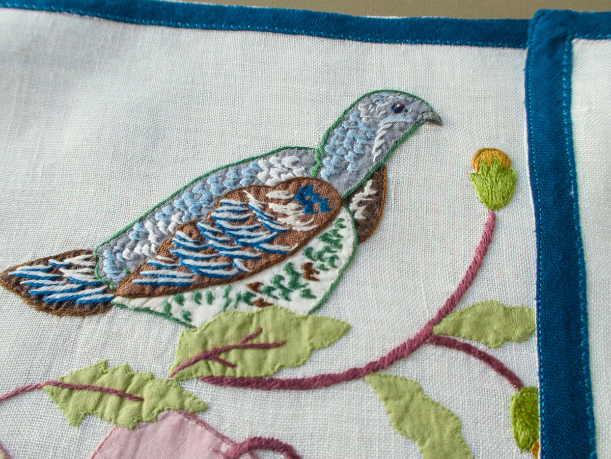 Close-up of a blue and brown embroidered bird on an Italian placemat, showing the stitching and blue border.