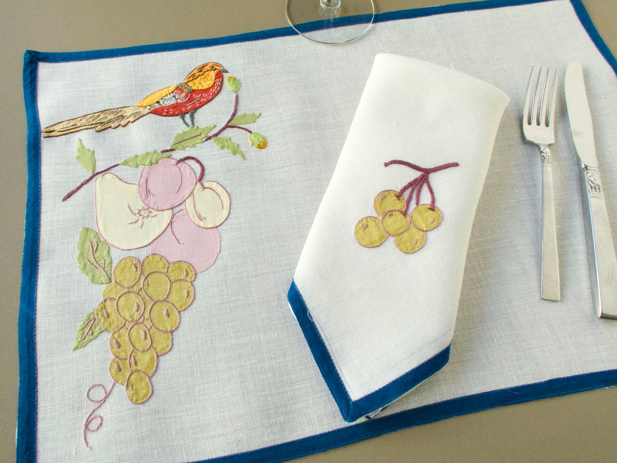 Italian placemat with a red and gold bird above fruit, set with a matching grape-embroidered napkin, silverware and a glass.