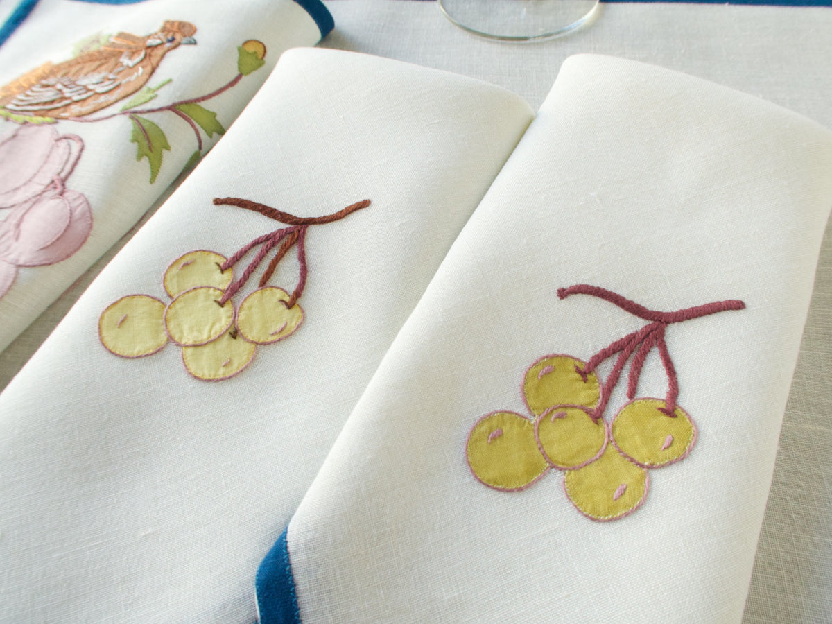 Two folded off-white napkins with embroidered grape clusters, laid over a matching placemat.