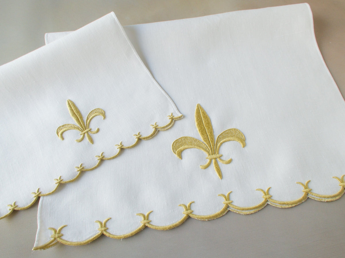 Marghab linen towels in the “Fleur de Lys” pattern, featuring raised gold embroidery with sculpted fleur-de-lys motifs and scalloped borders — guest and fingertip sizes shown together.