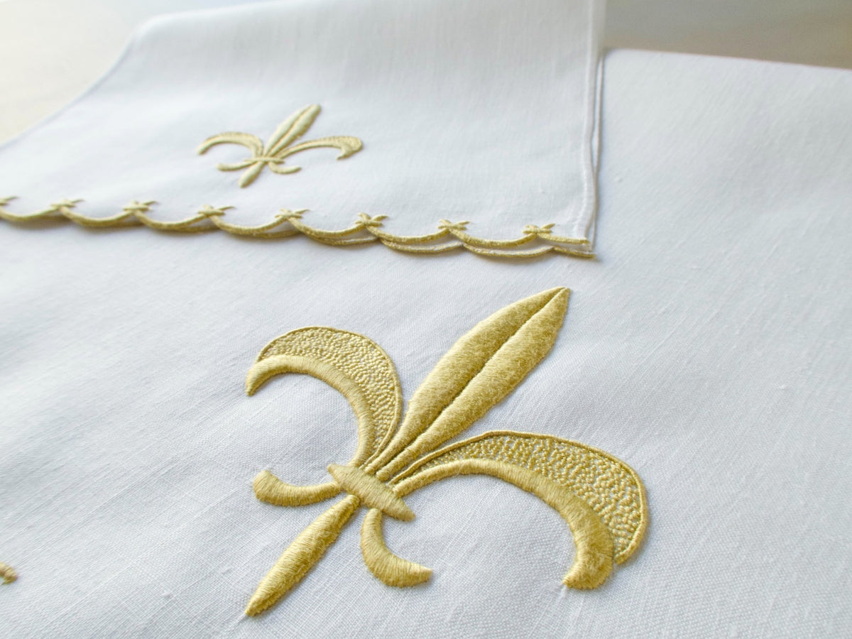 Marghab linen towels in the “Fleur de Lys” pattern, featuring raised gold embroidery with sculpted fleur-de-lys motifs and scalloped borders — guest and fingertip sizes shown together.