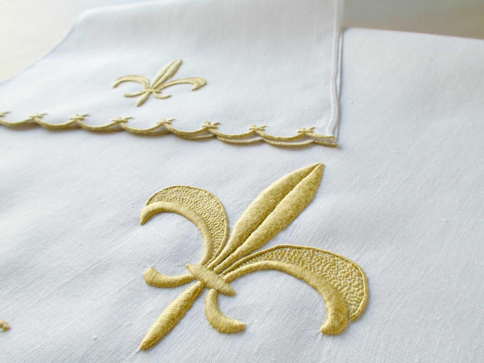 Marghab linen towels in the “Fleur de Lys” pattern, featuring raised gold embroidery with sculpted fleur-de-lys motifs and scalloped borders — guest and fingertip sizes shown together.