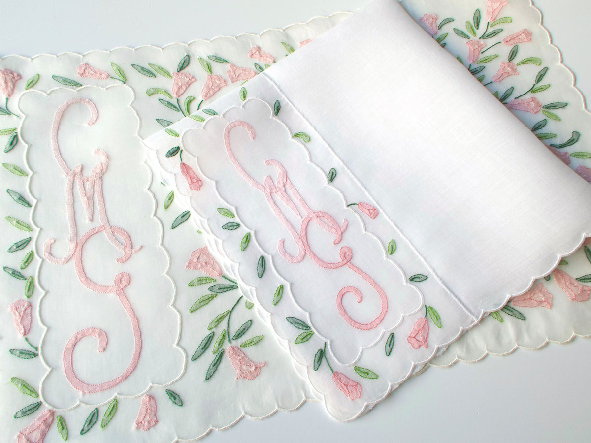Corner detail of Madeira placemat showing cursive pink monogram and scalloped edge alongside white linen napkin