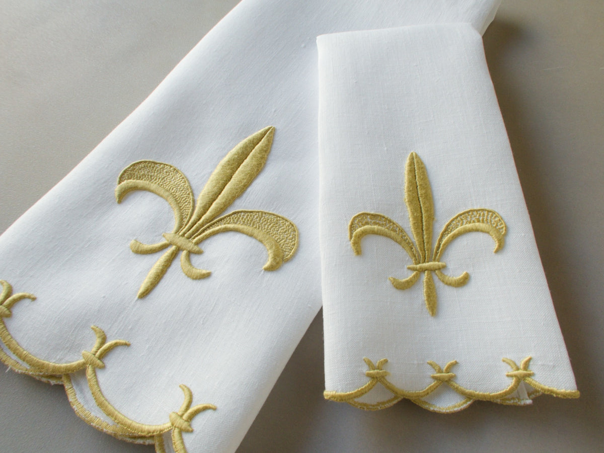 Marghab linen towels in the “Fleur de Lys” pattern, featuring raised gold embroidery with sculpted fleur-de-lys motifs and scalloped borders — guest and fingertip sizes shown together.