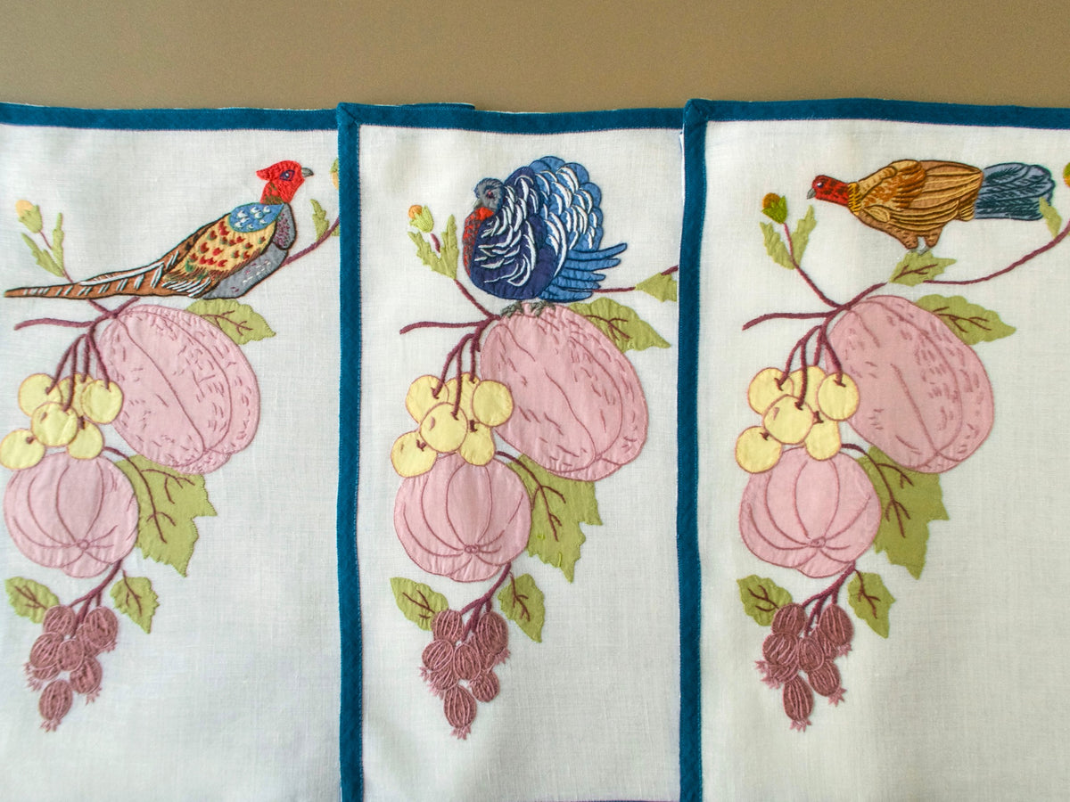Three placemats side by side, each with a different embroidered bird perched above cascading fruit.