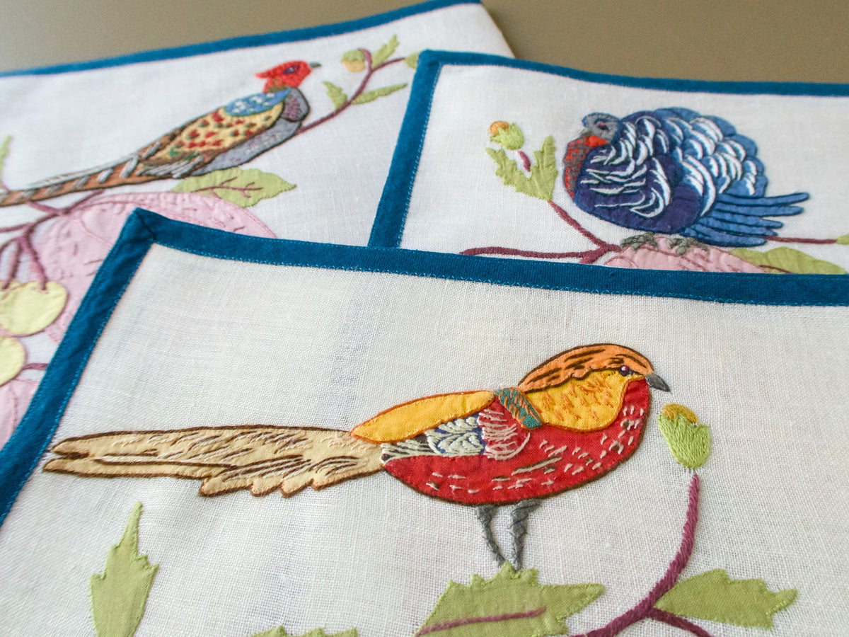 Three overlapping Italian placemats with embroidered birds above fruit, including a red and gold bird in front and a blue turkey behind.