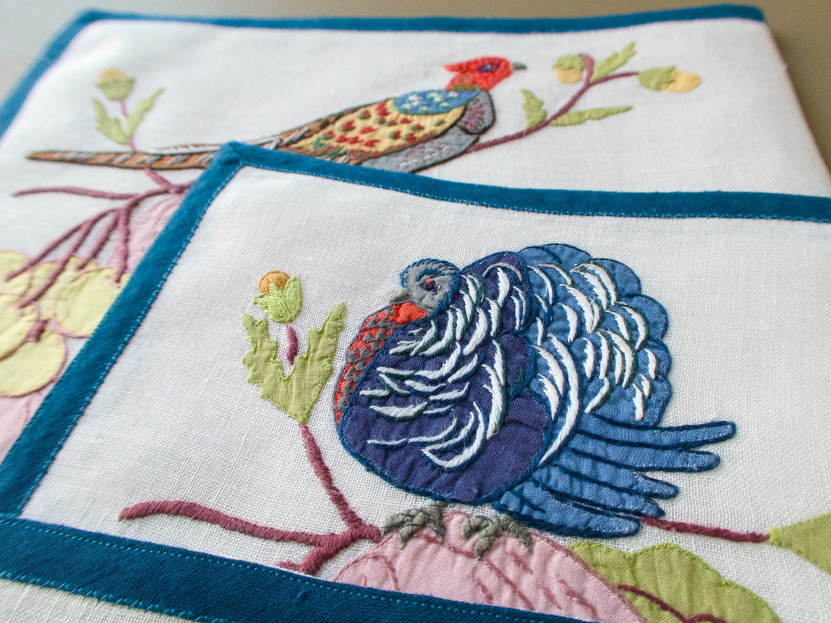 Close-up of a blue embroidered turkey on an off-white placemat with deep blue edging.