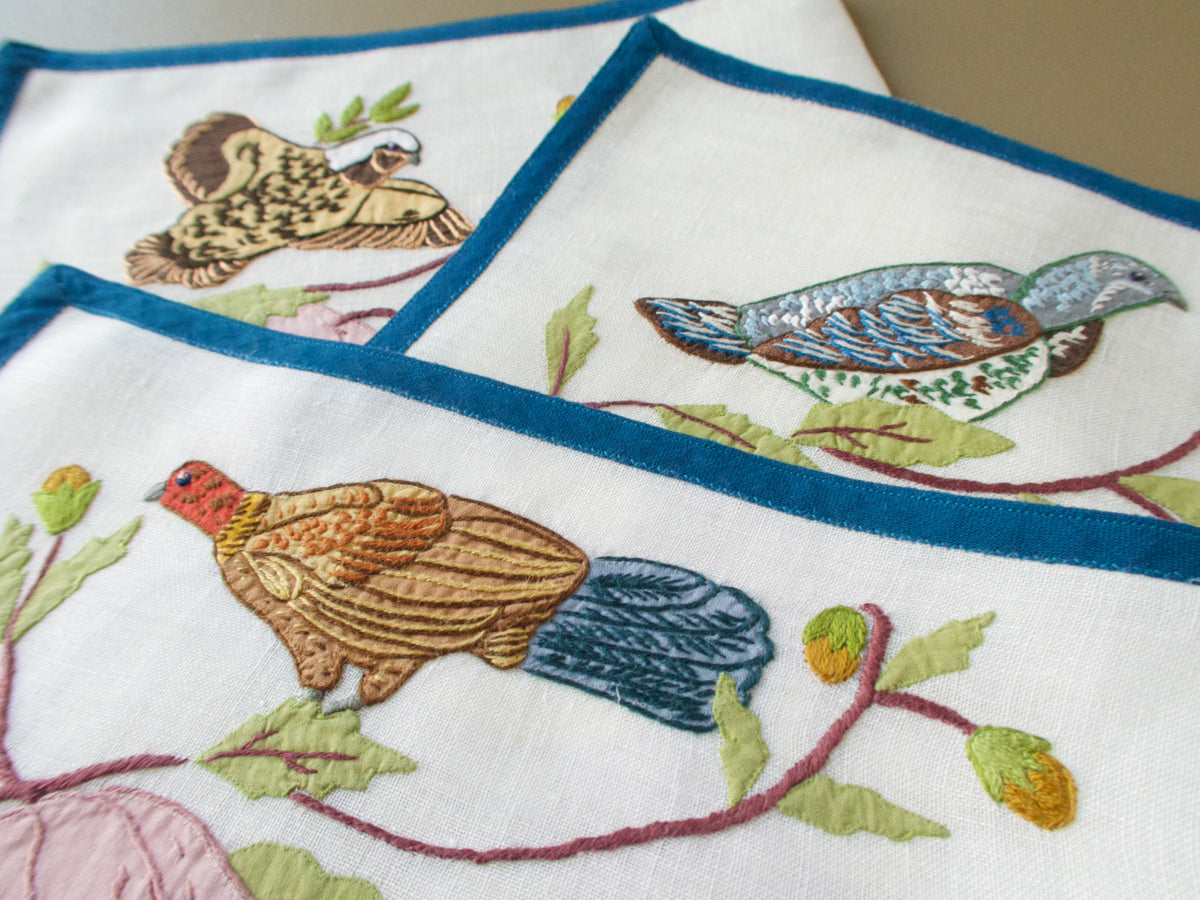 Three overlapping placemats showing the varied bird embroidery and deep blue linen borders.