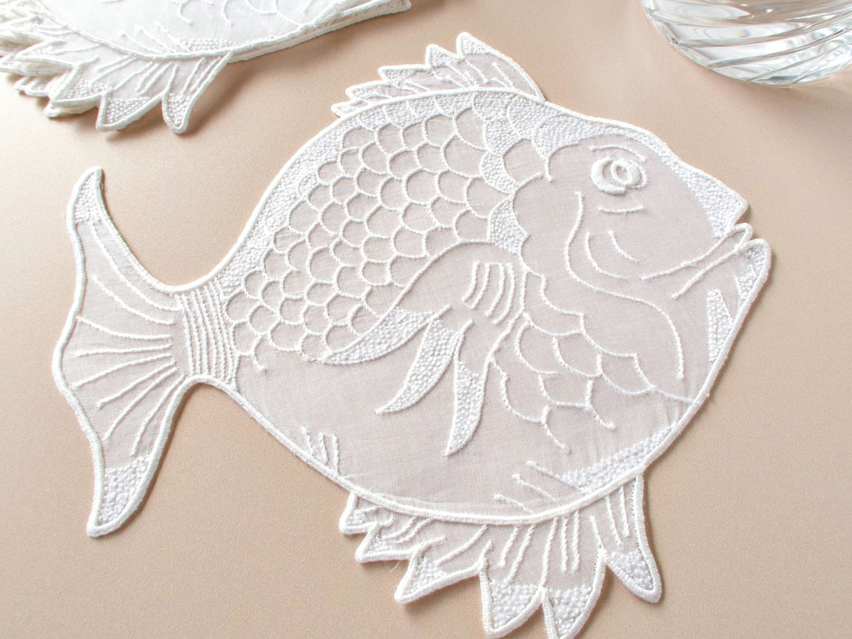 Fish-shaped vintage Madeira organdy cocktail napkin with finely embroidered scales, fins, and flowing lines on beige background