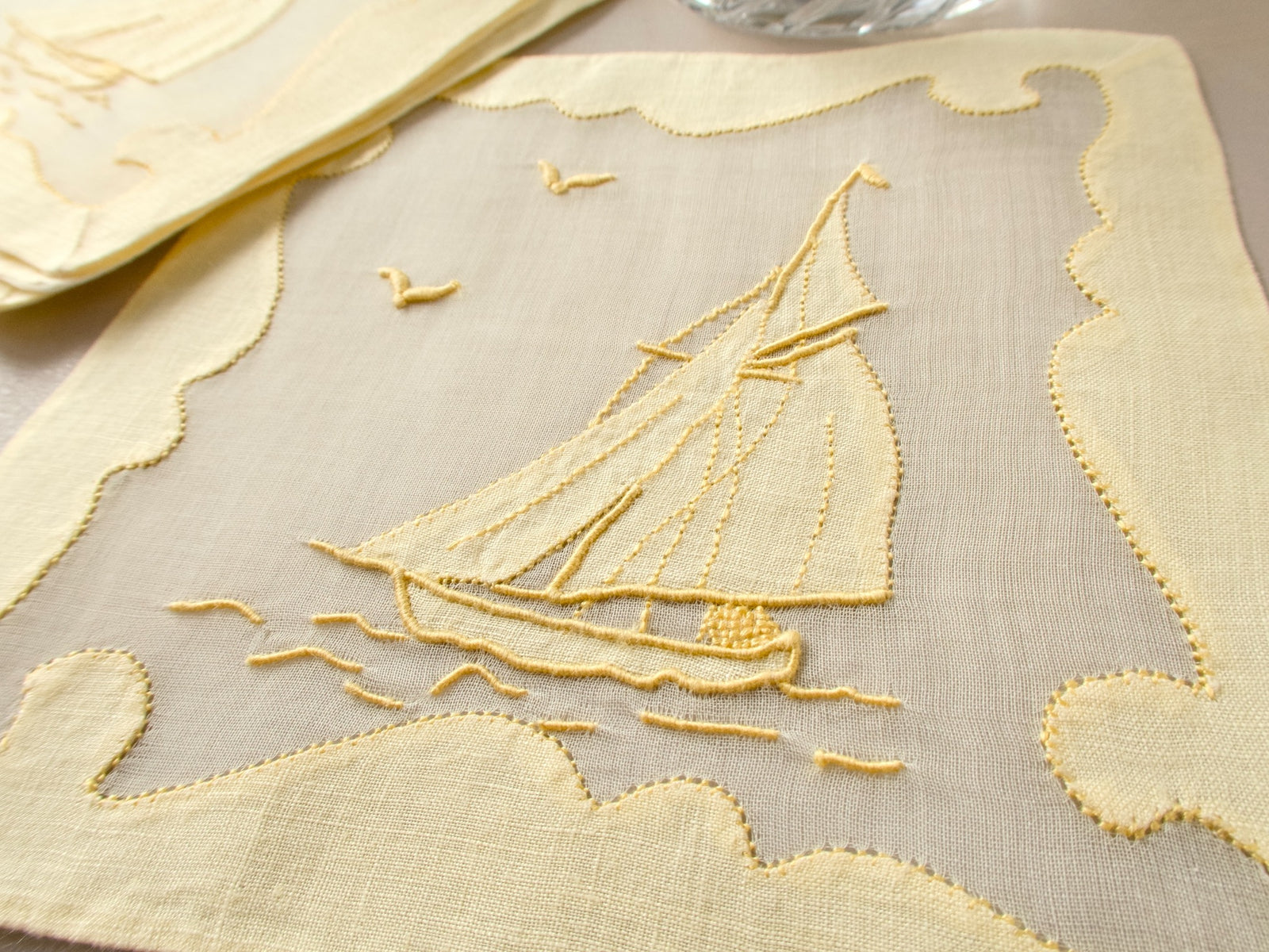Close-up of vintage Madeira cocktail napkin showing hand embroidered sailboat and ornate repliqué border in yellow linen​​​​​​​​​​​​​​​​