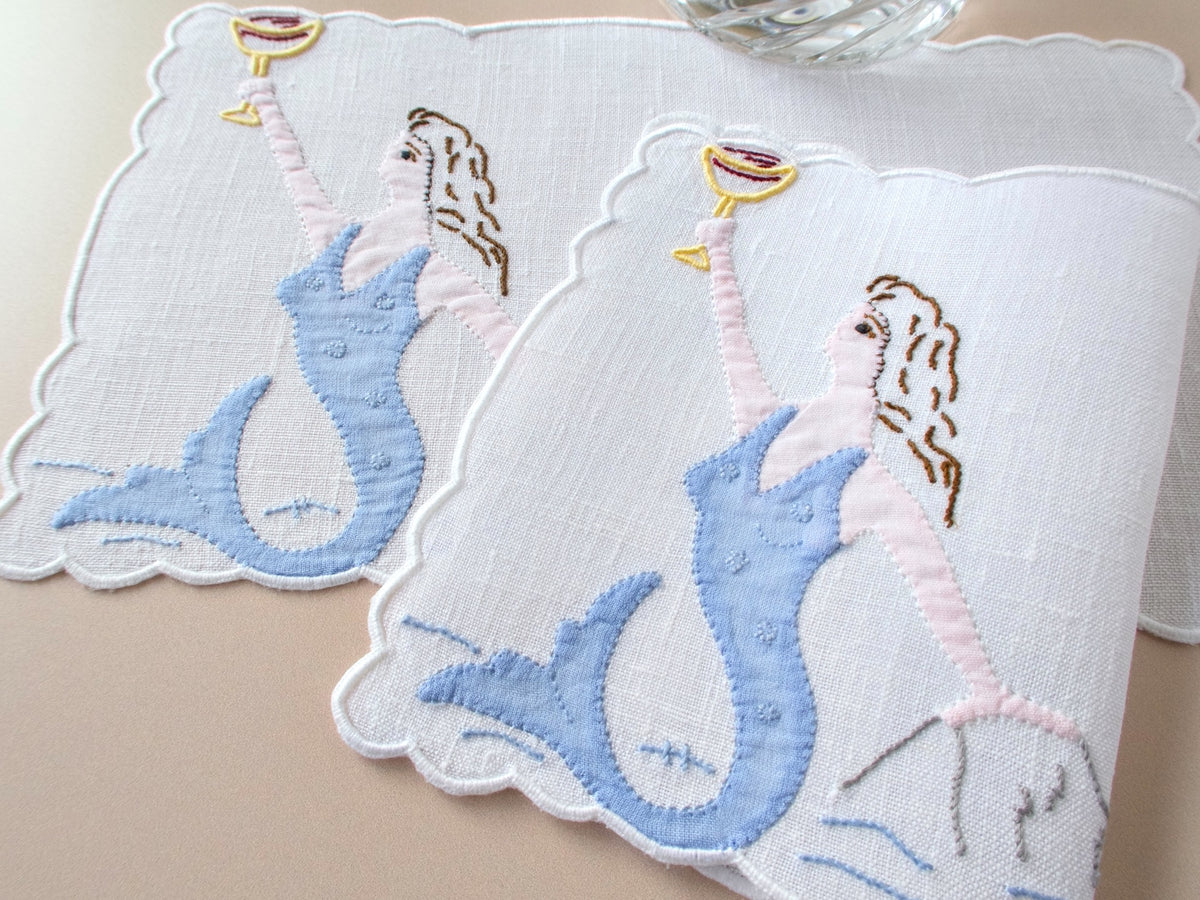 Pair of vintage Madeira linen cocktail napkins with blue appliqué mermaid raising golden goblet, scalloped border, on beige background
