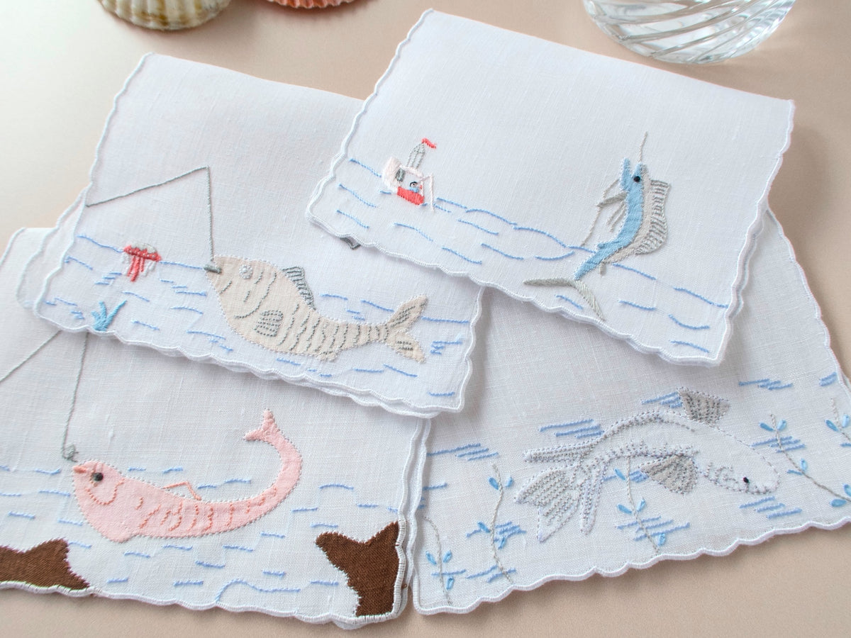 Set of four vintage Madeira linen cocktail napkins each with a different hand embroidered fish scene including marlin, whale, and fishing scenes on beige background