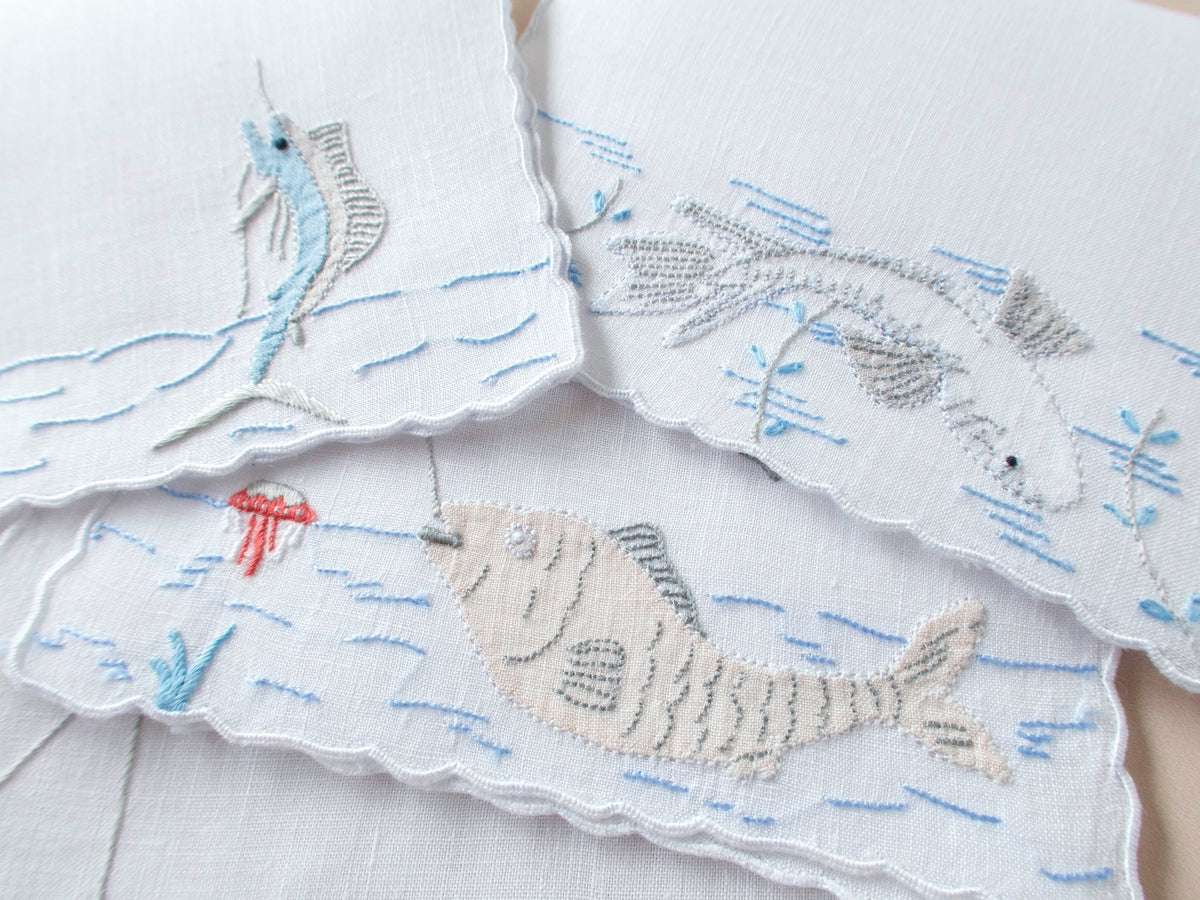 Close-up of vintage Madeira linen cocktail napkins showing hand embroidered fish appliqué and scalloped edges with blue wave detailing​​​​​​​​​​​​​​​​