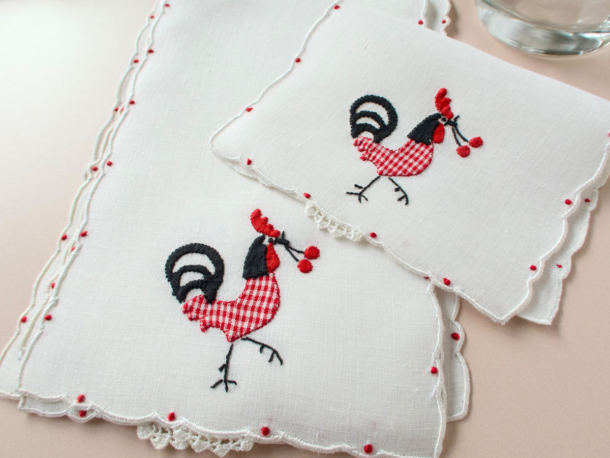 Two vintage Madeira linen cocktail napkins with red gingham rooster appliqué showing scalloped border and red dot accent detail on beige background​​​​​​​​​​​​​​​​