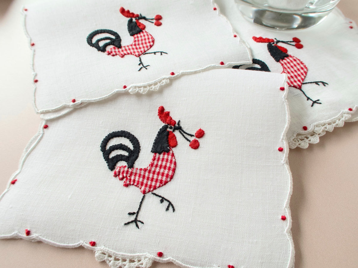 Vintage Madeira linen cocktail napkins with red gingham rooster appliqué and red dot scalloped border, stacked on beige background with glass for scale