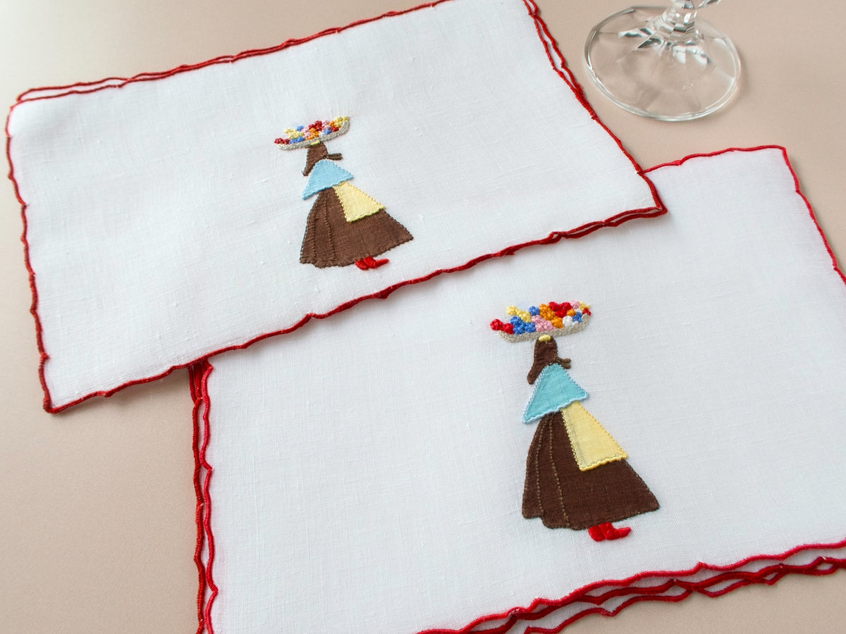 Two vintage Marghab Flower Woman linen cocktail napkins with red scalloped border showing appliqué figure balancing flower basket on beige background