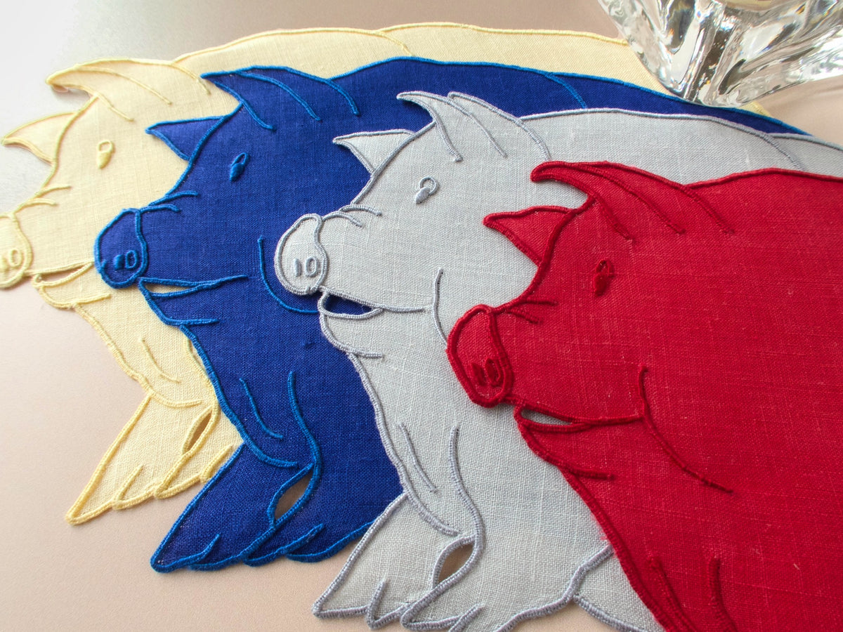 Close-up of four pig-shaped vintage Madeira cocktail napkins showing expressive pig faces and embroidered detail in blue, gray, yellow, and red​​​​​​​​​​​​​​​​