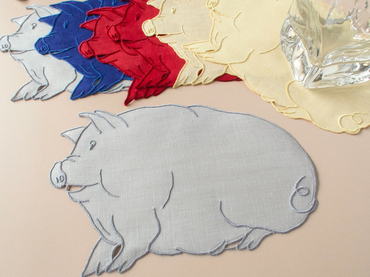 Pig-shaped vintage Madeira linen cocktail napkin in gray with full set of eight pigs in all four colors and glass for scale on beige background