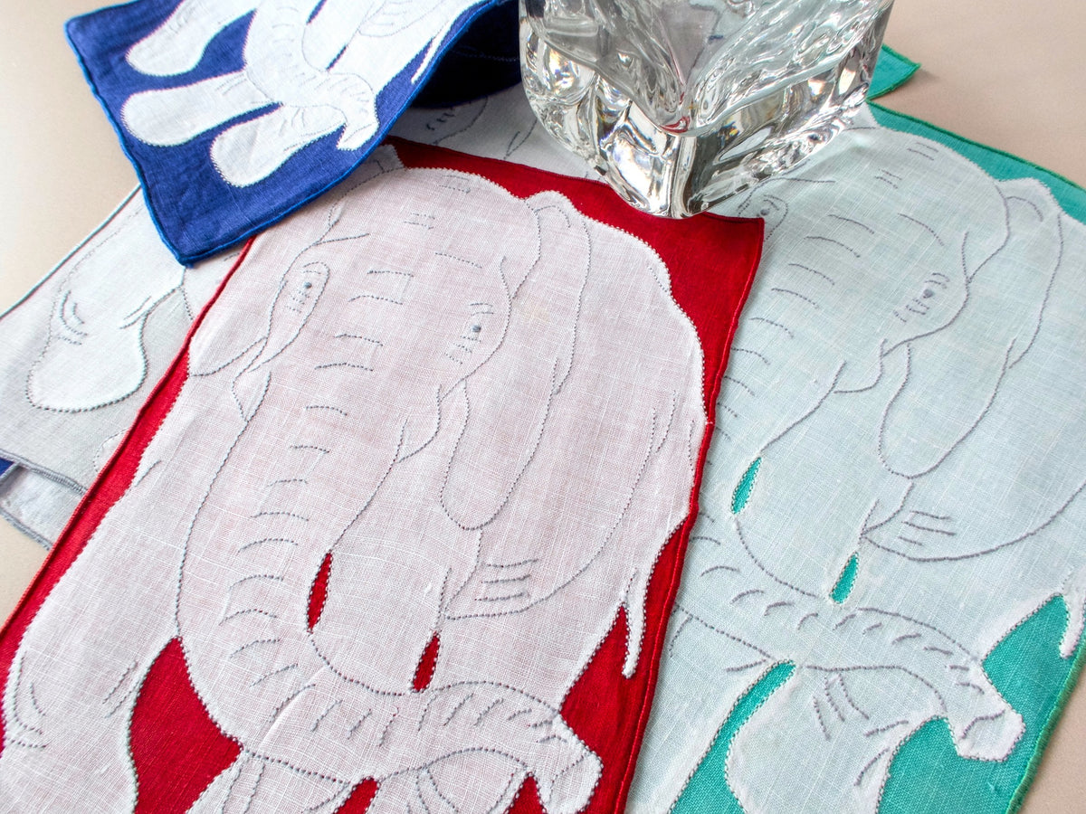 Three vintage Madeira elephant cocktail napkins in red, teal, and blue with glass for scale showing white linen appliqué detail​​​​​​​​​​​​​​​​