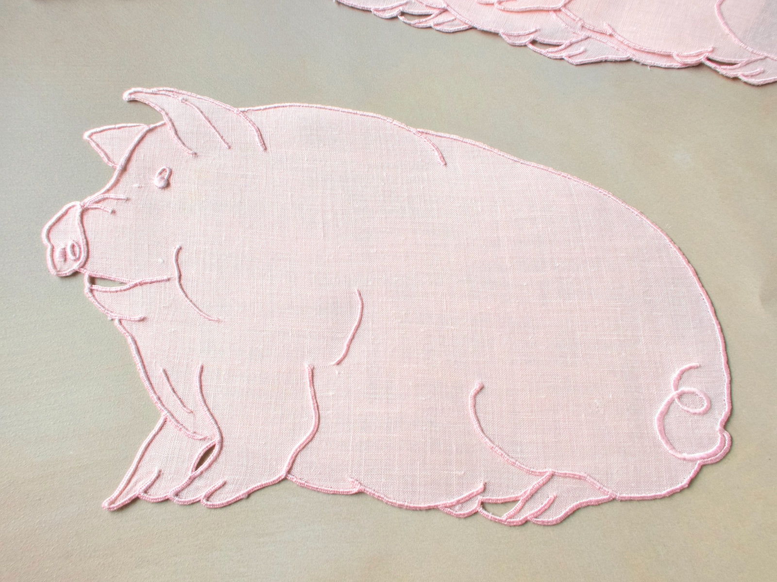 Close-up of a single pink Irish linen cocktail napkin cut and embroidered in the shape of a plump pig with curly tail, Madeira work.​​​​​​​​​​​​​​​​