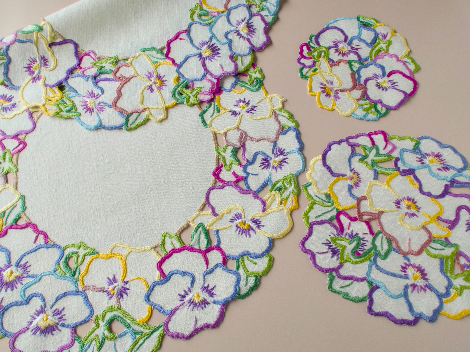 Vintage hand-embroidered pansy placemat set on beige background, multicolor thread in purple, yellow, blue, pink and green on white linen.