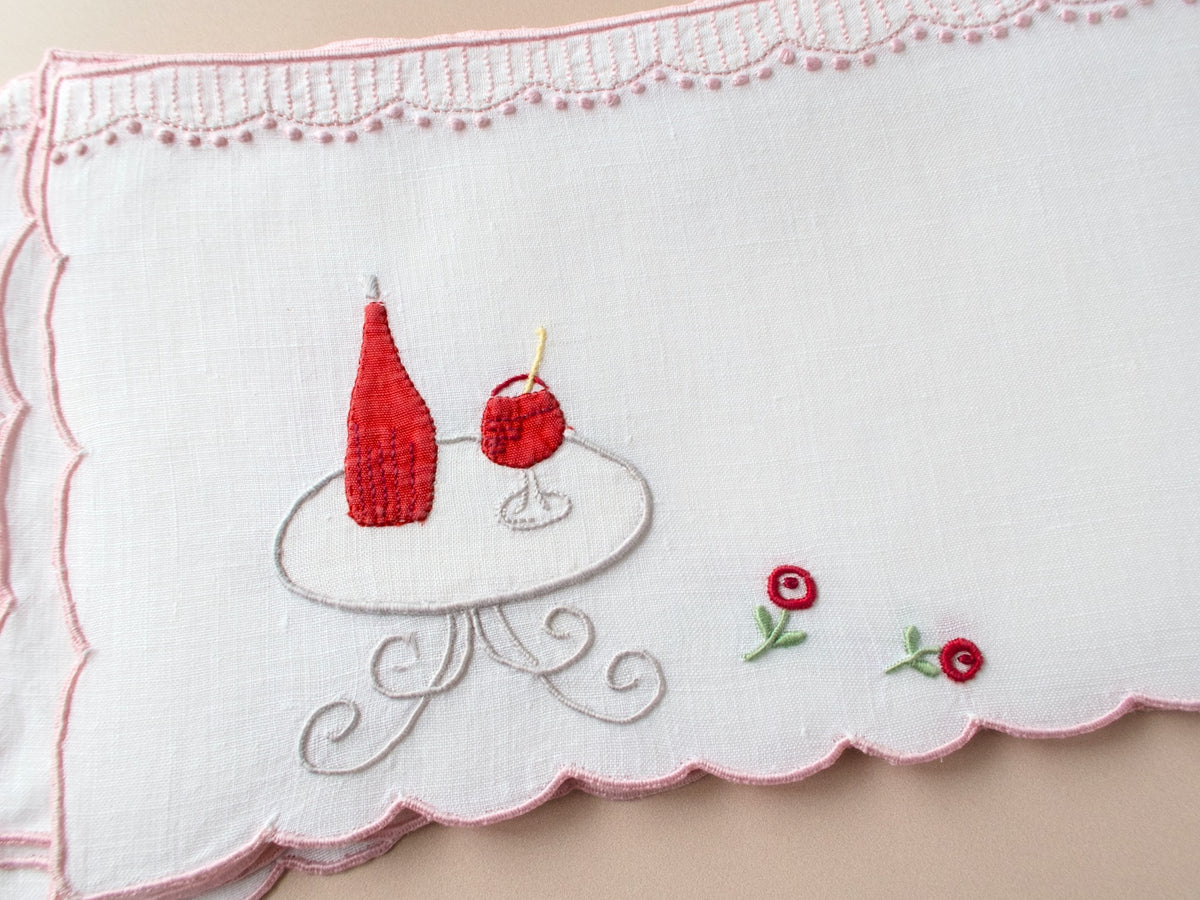 Vintage Madeira cocktail napkins on beige background, red wine bottle and glass embroidered on scrollwork café table with roses, pink scalloped border.