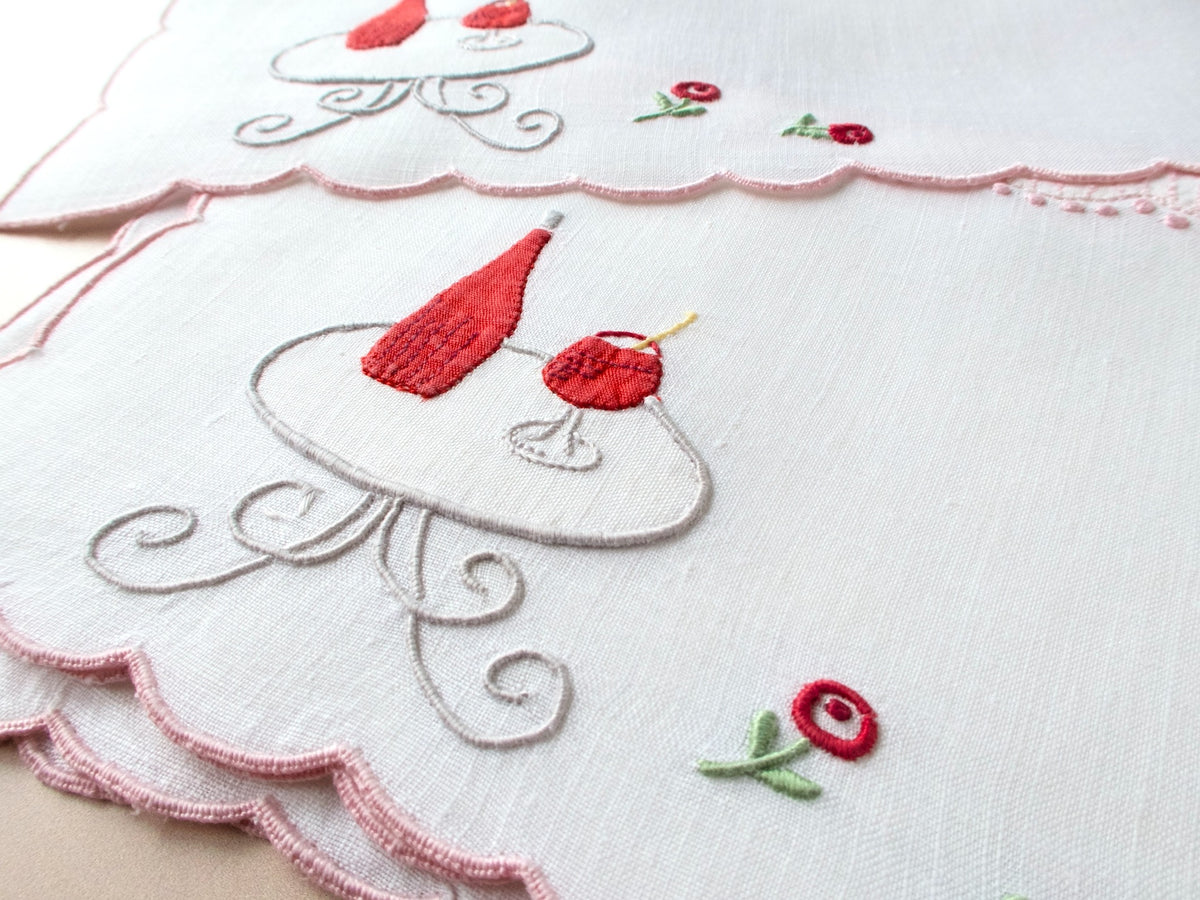 Close-up of vintage Madeira cocktail napkin showing red embroidered wine bottle and glass on scrollwork table with rose detail and pink scalloped border.​​​​​​​​​​​​​​​​