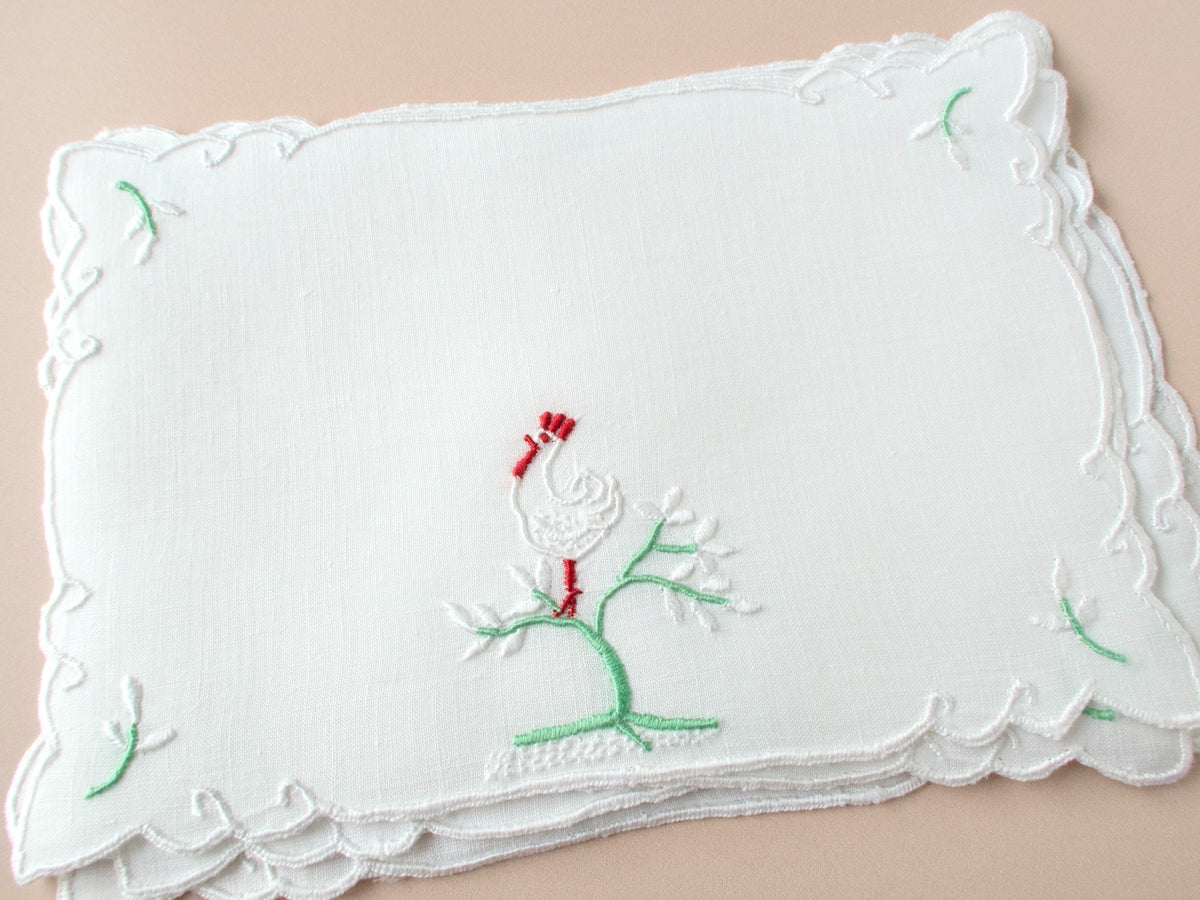 Vintage Madeira cocktail napkins on beige background, white embroidered rooster on green flowering tree with red comb, white scalloped border.