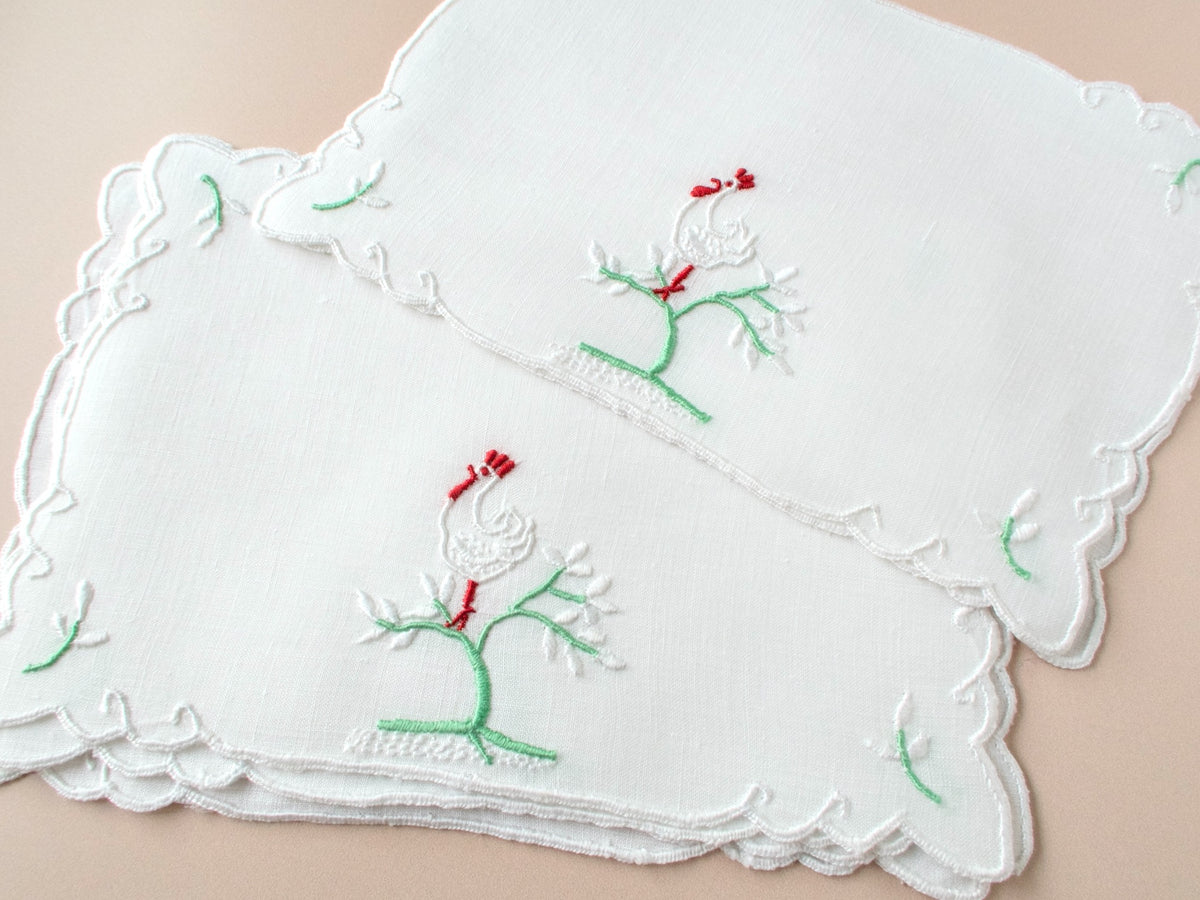 Two vintage Madeira cocktail napkins fanned on beige background showing rooster on flowering tree embroidery and white scalloped edge.