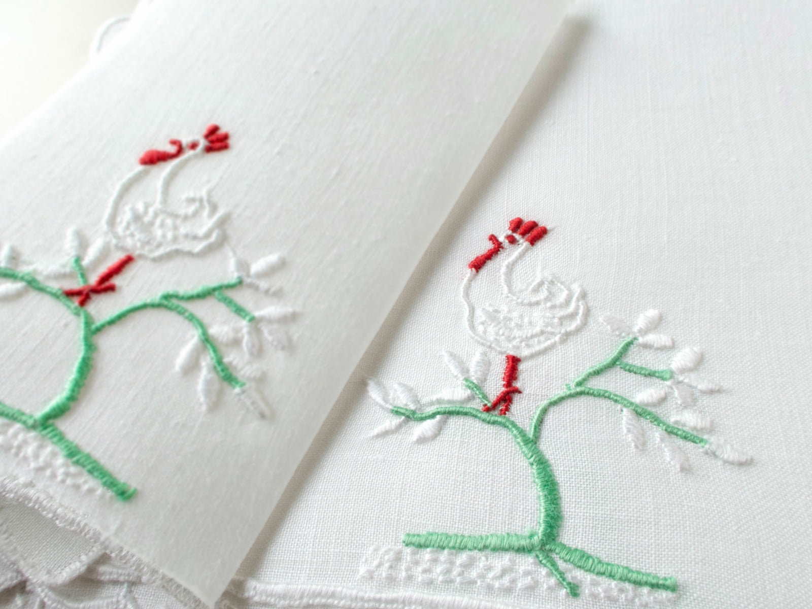 Close-up of vintage Madeira cocktail napkins showing white satin stitch rooster with red comb on green embroidered tree branches.​​​​​​​​​​​​​​​​