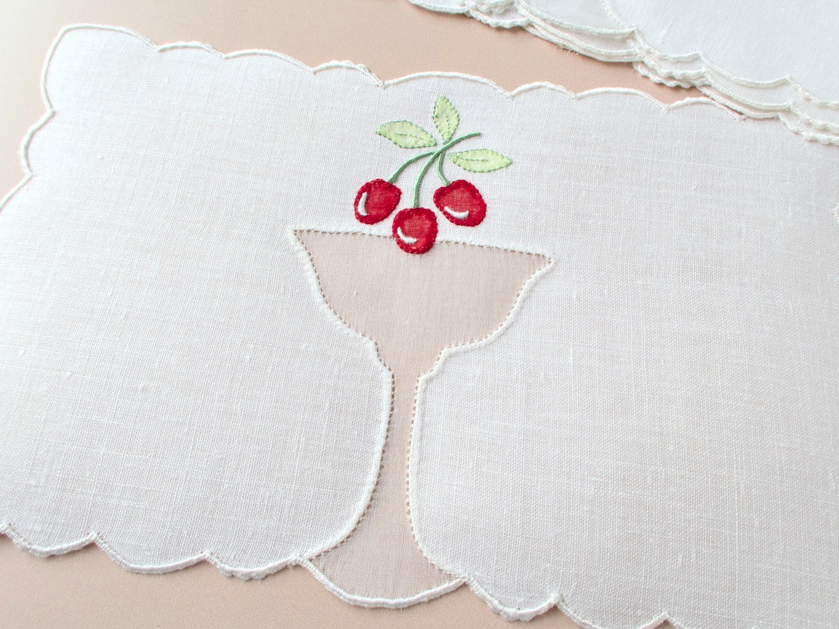 Single vintage Madeira cocktail napkin on beige background showing full design — organdy cocktail goblet inset with red cherry appliqué and scalloped border.