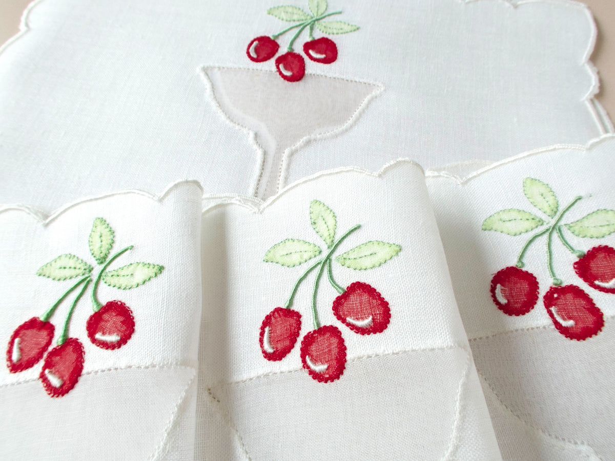 Close-up of vintage Madeira cocktail napkin showing luminous sheer organdy cherry appliqué with hand embroidered stems, leaves and scalloped white border.