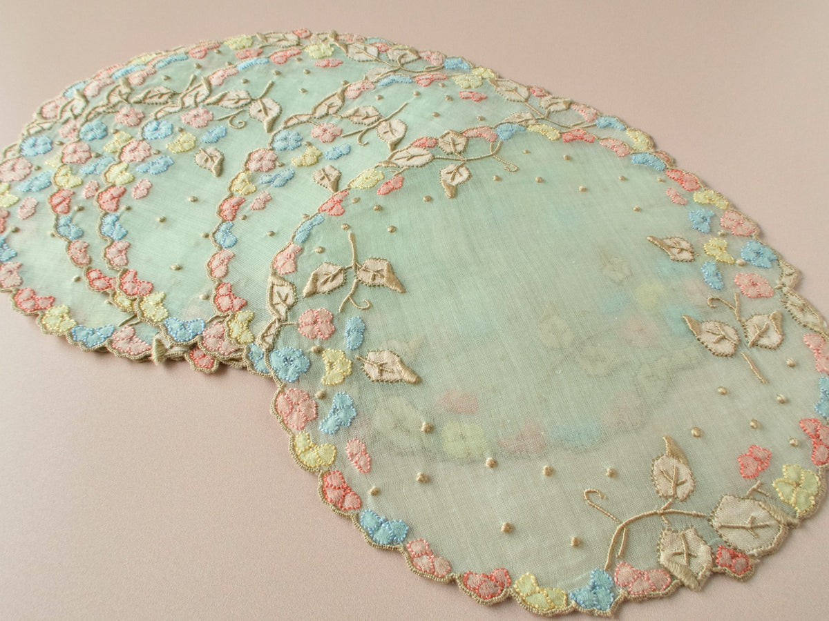 Vintage Madeira organdy cocktail rounds on beige background, pale green sheer organdy with multicolor pink blue and yellow flower border and embroidered vines with granitos.