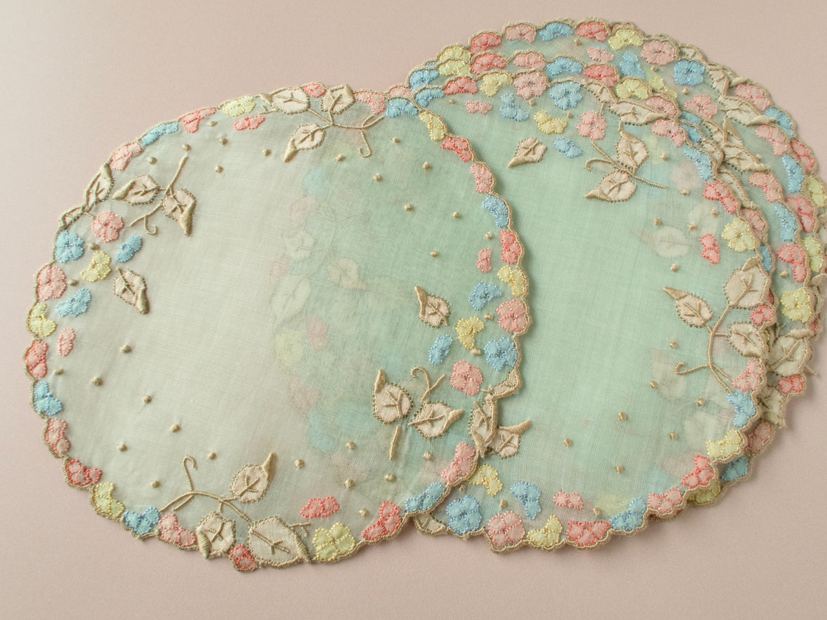 Stacked vintage Madeira pale green organdy cocktail rounds showing full design — multicolor flower garland border, shadow work leaves and embroidered vine detail.