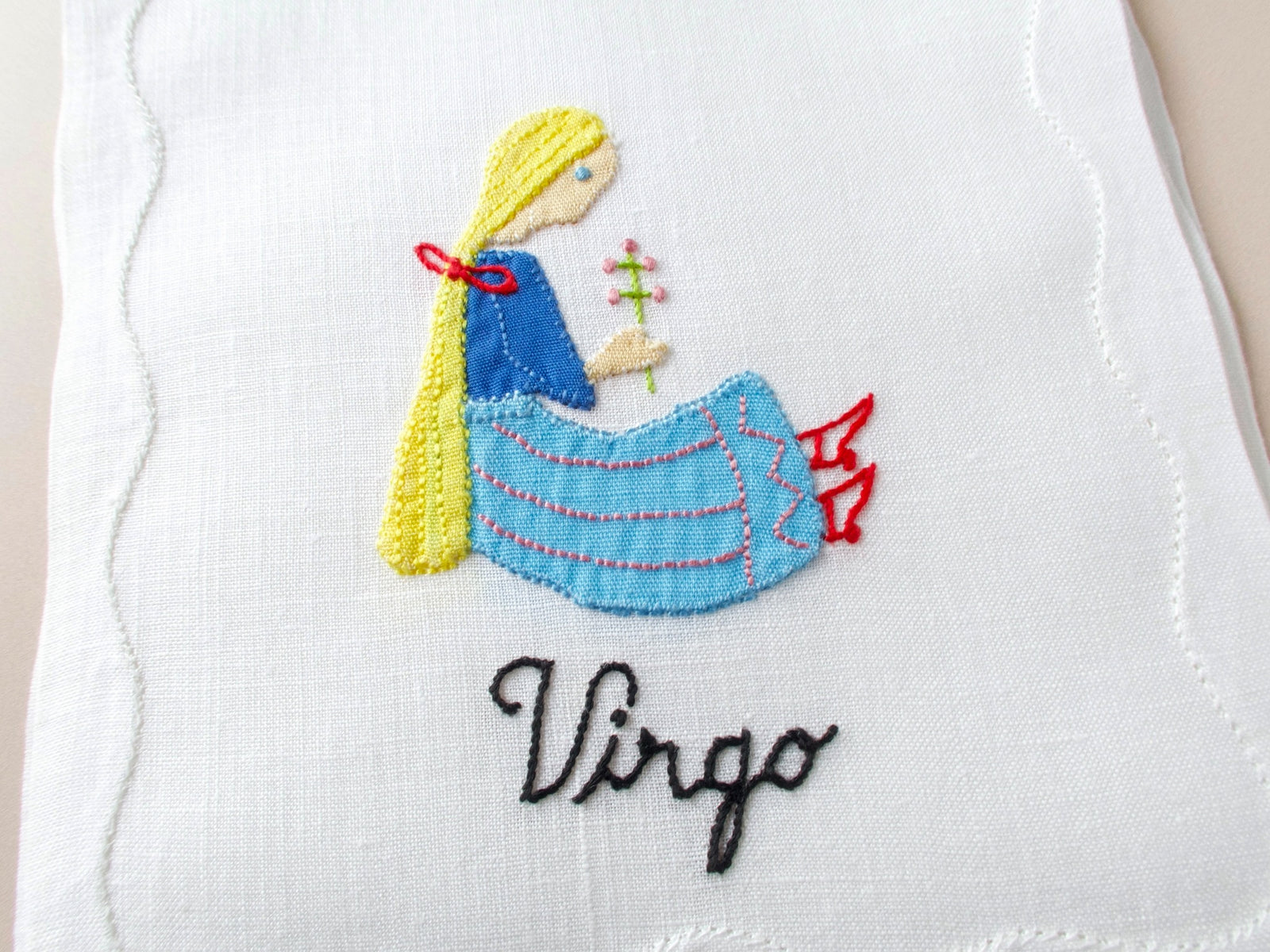 Close-up of vintage Madeira cocktail napkin showing detailed appliqué Virgo maiden in blue dress holding flower sprig with embroidered script in black thread on white linen.​​​​​​​​​​​​​​​​