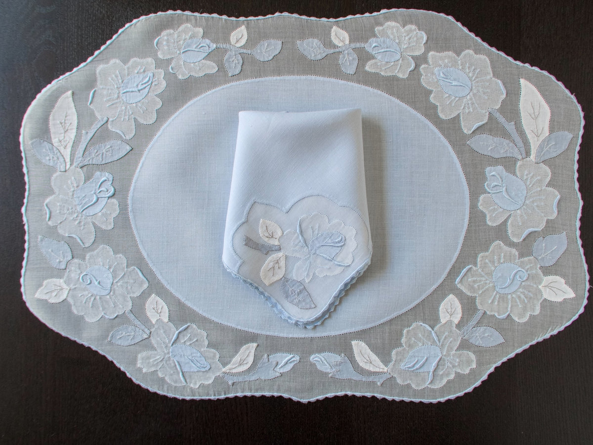 Vintage Madeira pale blue organdy placemat set for 8 showing complete oval shape with appliqué rose border, folded napkin with matching motif,  dark background.