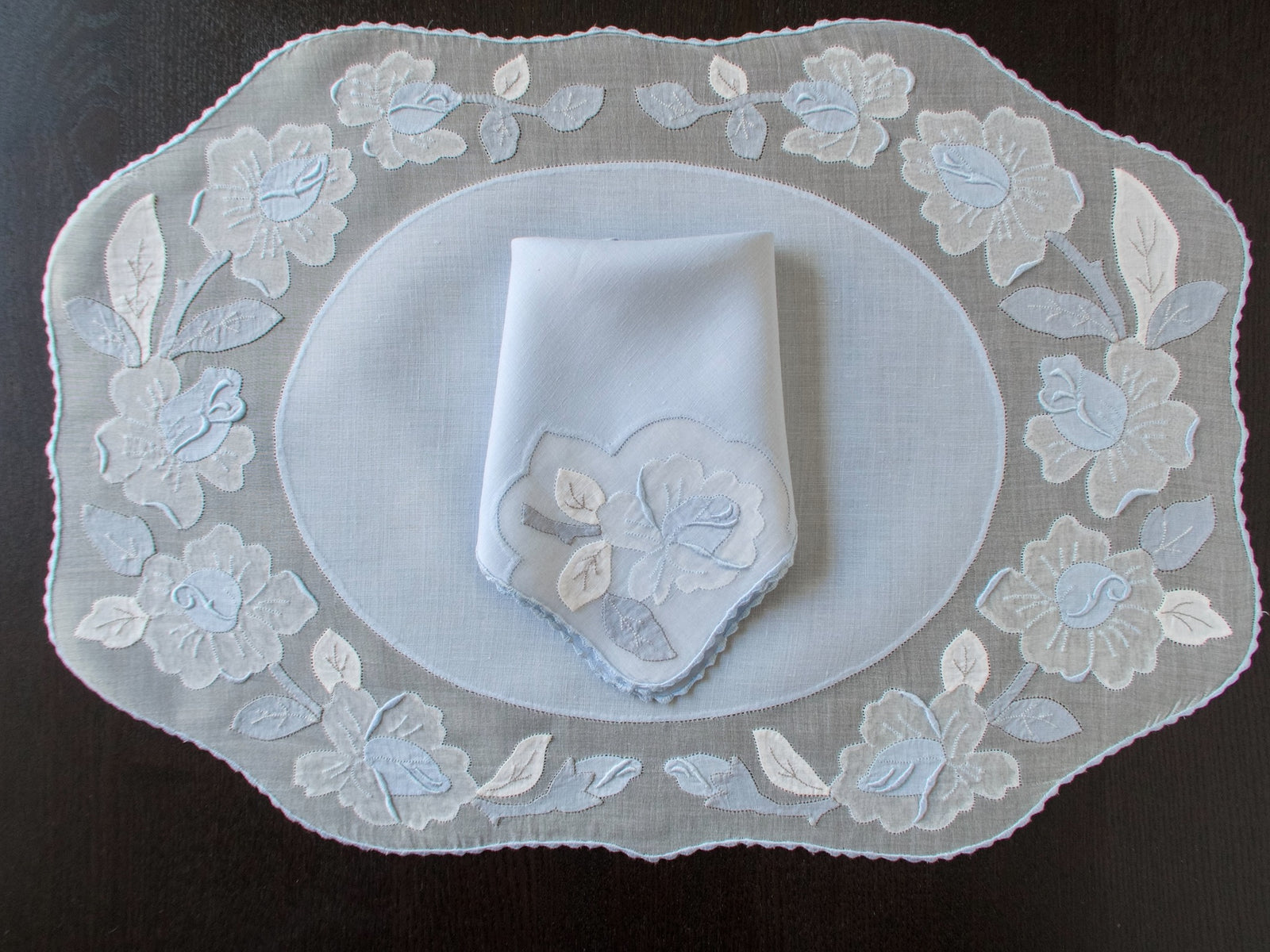 Vintage Madeira pale blue organdy placemat set for 8 showing complete oval shape with appliqué rose border, folded napkin with matching motif,  dark background.