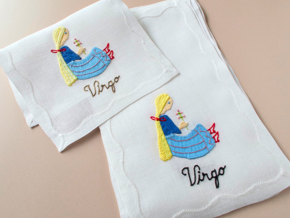 Vintage Madeira cocktail napkins on beige background showing two napkins with appliqué Virgo maiden in blue dress and yellow hair with embroidered Virgo script below.