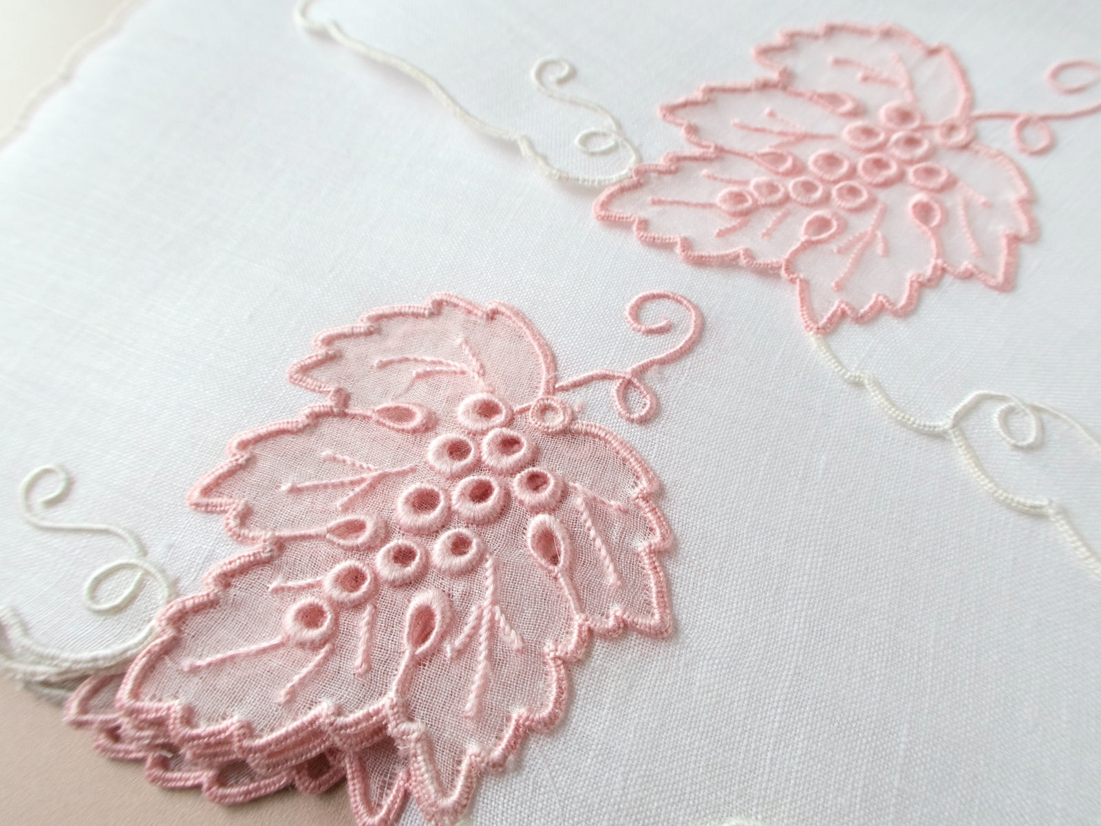 Close-up of vintage Madeira cocktail napkin showing sheer blush pink organdy grape leaf with raised satin stitch grape clusters and white scrolling vine detail.