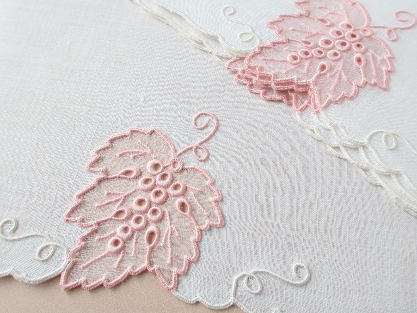 Detail shot of vintage Madeira cocktail napkin blush pink organdy grape leaf and raised grape cluster embroidery on white linen with scalloped border.​​​​​​​​​​​​​​​​