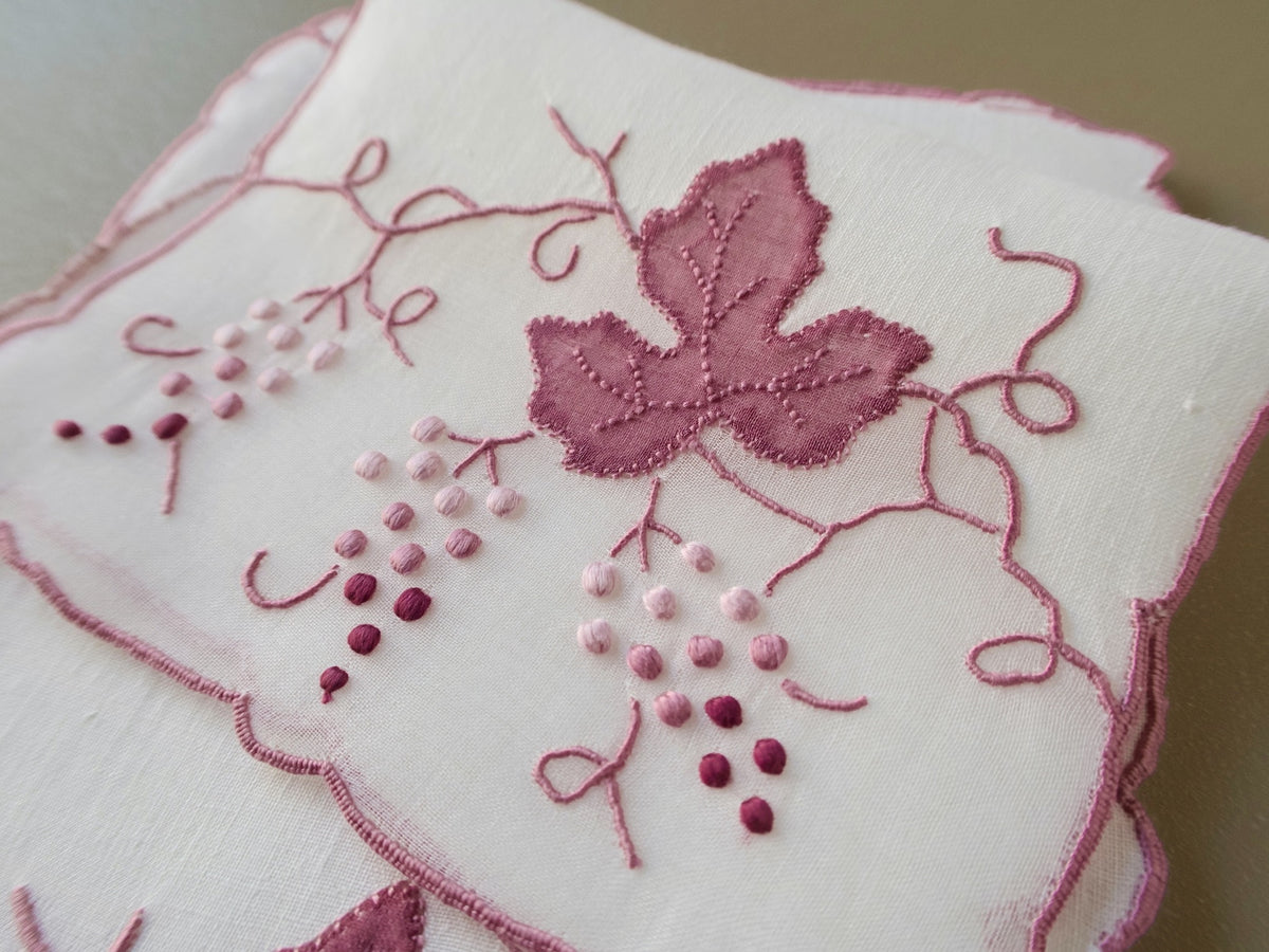 Closeup of a vintage Madeira cocktail napkin on beige background raised pink grape clusters and leaf appliqué with scrolling vines.