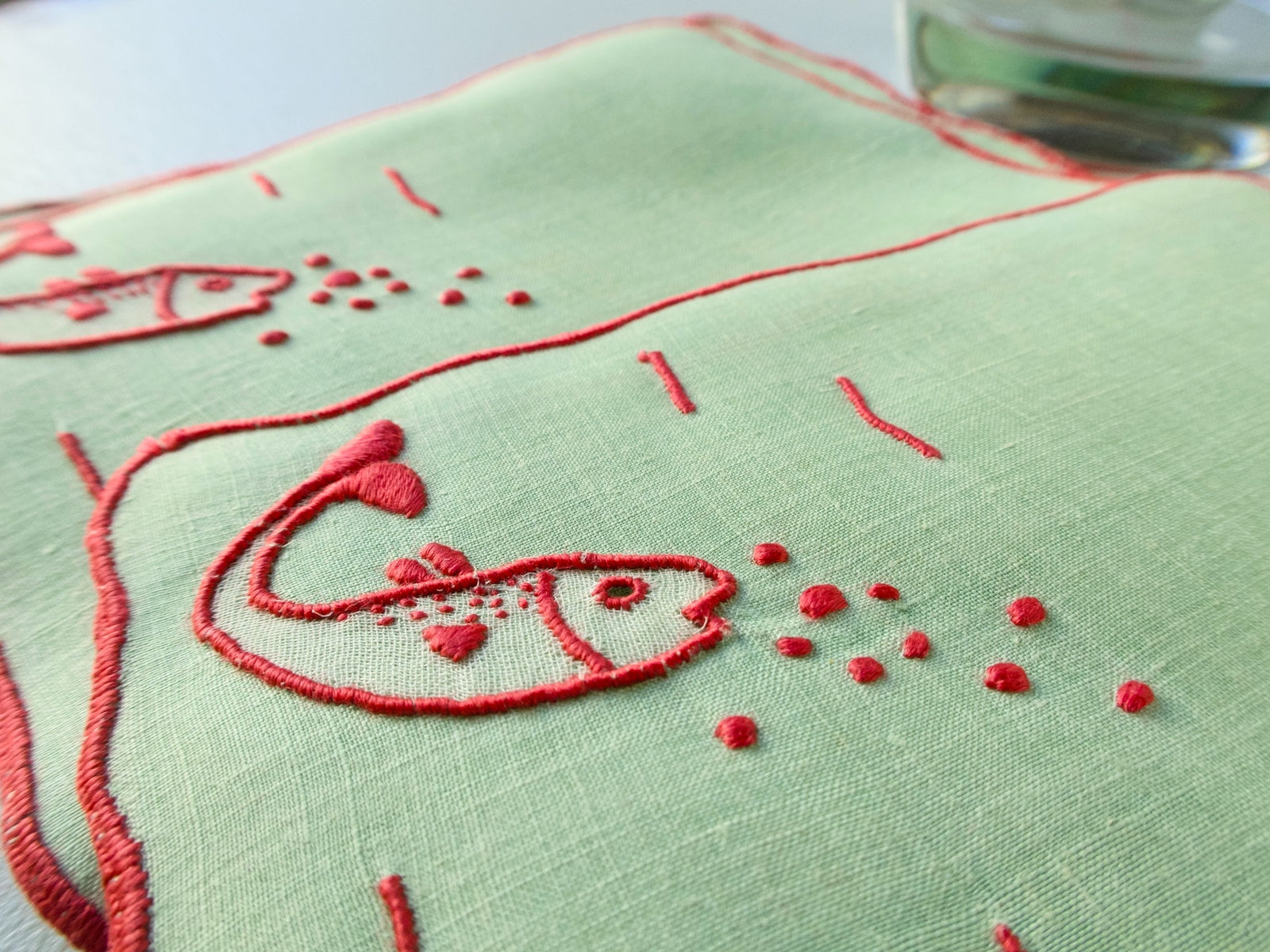 Close view of Marghab “Fish” pattern — red fish with bubbles hand embroidered on soft green Madeira Irish linen cocktail napkin.​​​​​​​​​​​​​​​​