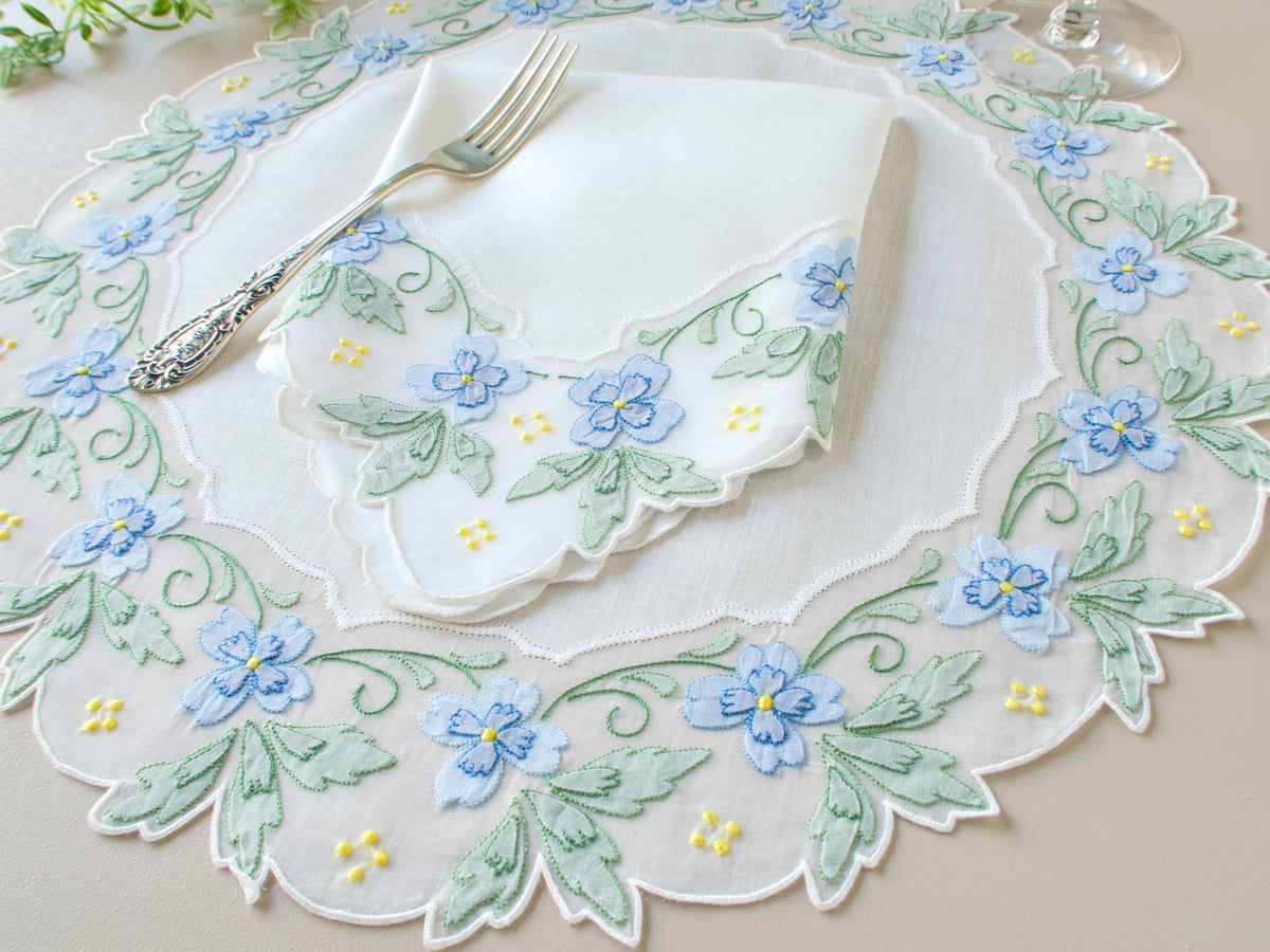 Vintage Madeira round placemat with blue and green shadow work flowers and leaves on scalloped organdy border, styled with linen napkin and silver fork
