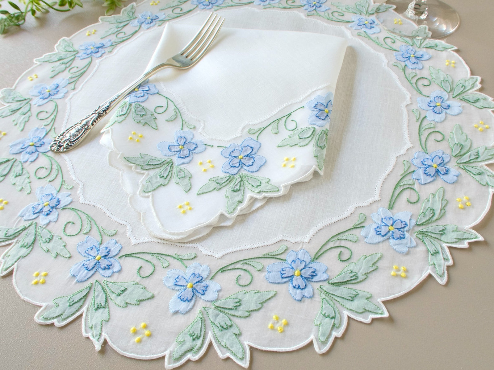 Vintage Madeira round placemat with blue and green shadow work flowers and leaves on scalloped organdy border, styled with linen napkin and silver fork