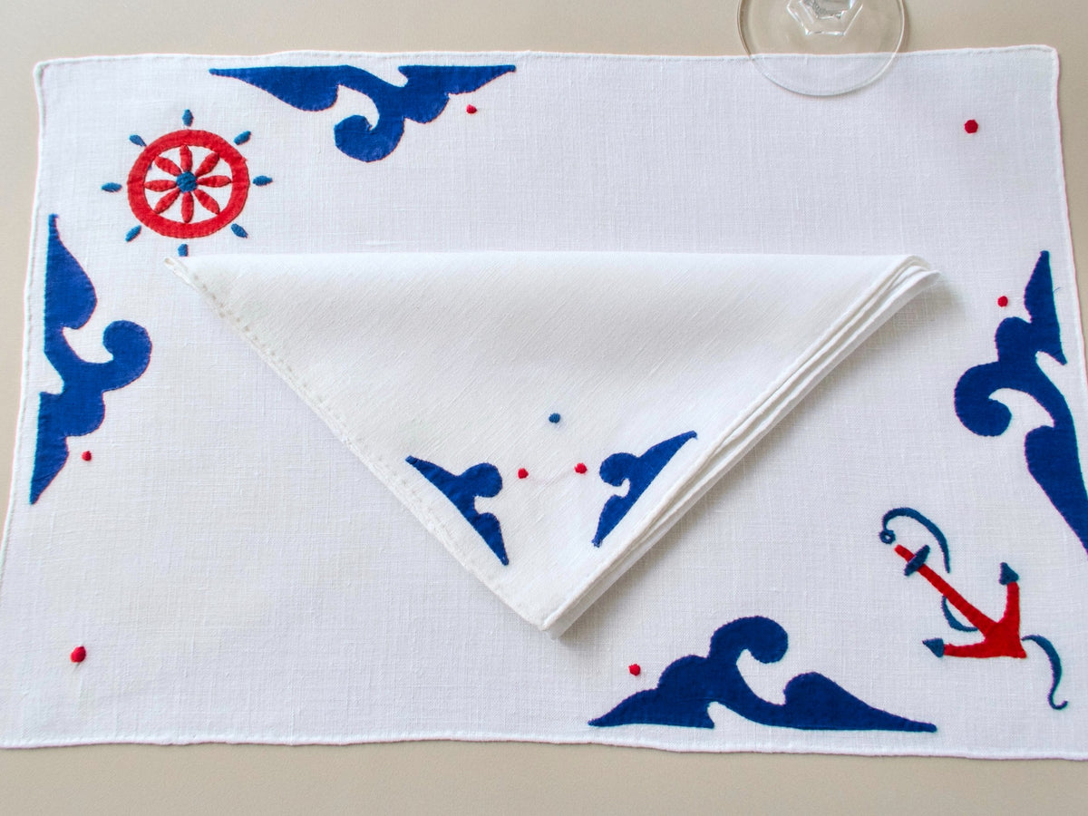 Overhead view of vintage Italian nautical embroidery and appliqué — anchor, ship’s wheel and wave motifs in navy and red on white linen.